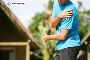 A man wearing a blue shirt holds his shoulder and elbow, suggesting discomfort or pain, with a house and trees in the background.