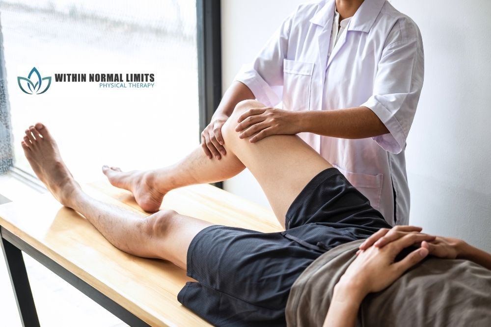 A physical therapist in a white coat examines a patient's leg on an exam table in a clinic with "Within Normal Limits Physical Therapy" logo on the wall.
