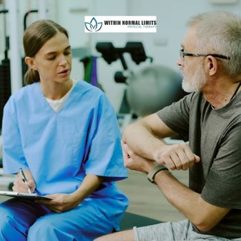 A physical therapist in blue scrubs consults with an older man about his elbow in a clinic setting.