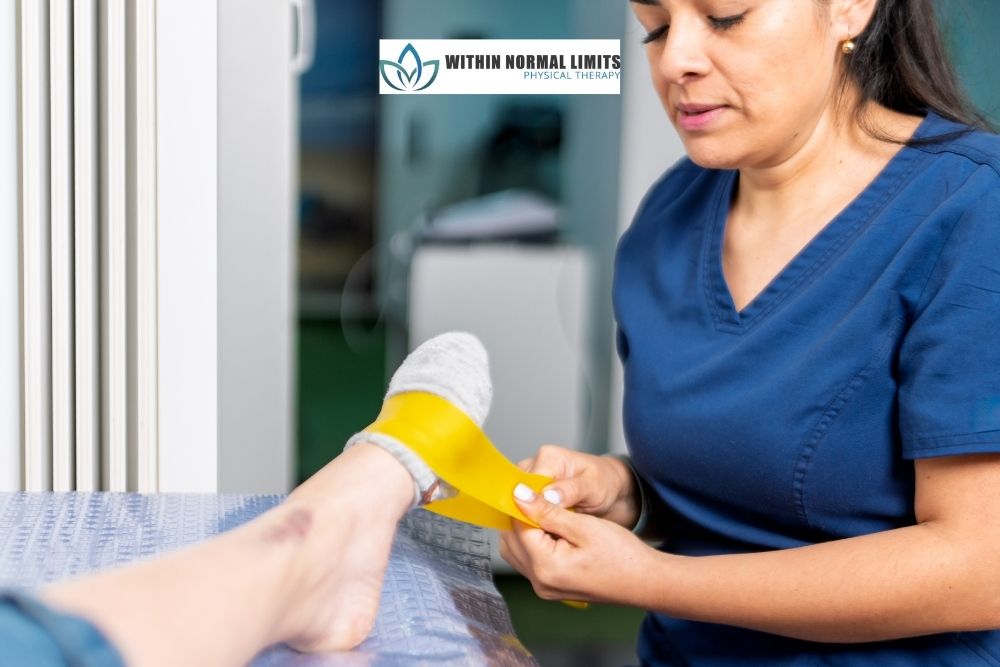 A physical therapist in blue scrubs helps a patient with a socked foot perform a resistance band exercise at a clinic.